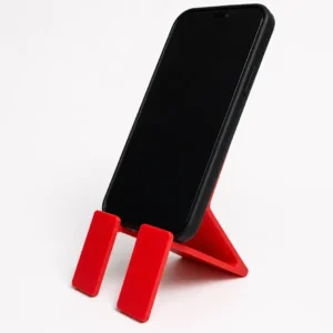 oporte-celular-bioplastico-rojo-uso-escritorio Teléfono celular negro con funda colocado en posición vertical sobre un soporte de bioplástico rojo, con fondo blanco.