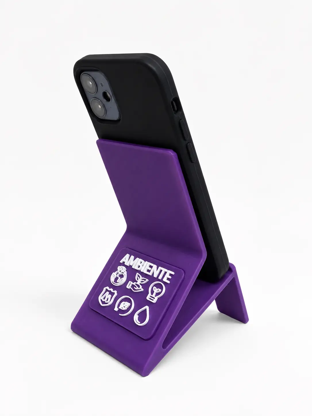 Teléfono celular negro colocado de espaldas sobre un soporte de bioplástico violeta con la inscripción AMBIENTE e íconos ecológicos en blanco, sobre fondo blanco.